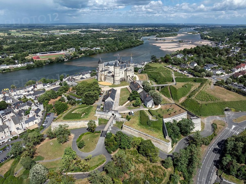 Aerial view of the historic Château de Saumur with its distinctive round corner towers and bright facades, surrounded by well-kept green spaces under a slightly cloudy summer sky, with the Loire River, Maine-et-Loire, Pays de la Loire, France