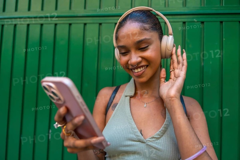 Young woman smiling while listening to music with wireless headphones, engaging with her smartphone outdoors against a vibrant green background