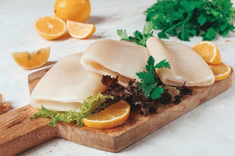 Fresh squid prepared for cooking, surrounded by herbs and lemon, placed on a wooden chopping board in a bright kitchen setting