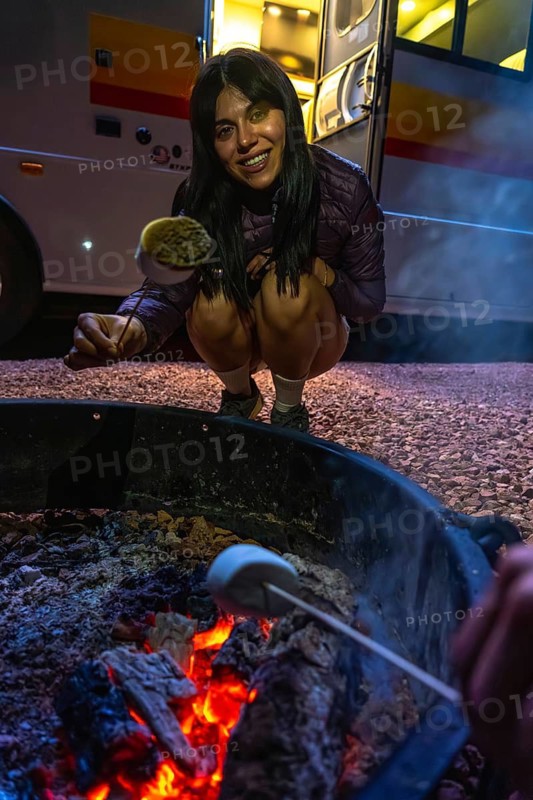 Young woman smiles while roasting marshmallows over a glowing portable campfire pit beside an rv at dusk, enjoying a relaxed evening of camping, s'mores and outdoor warmth