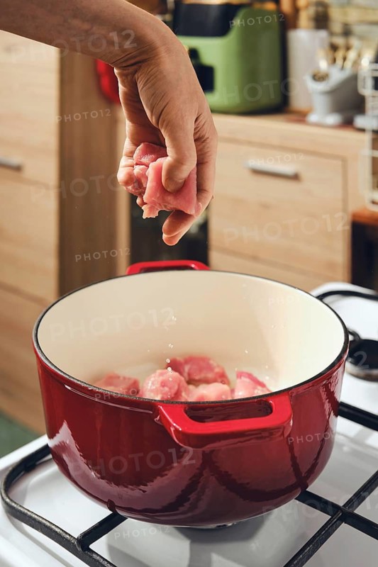 Hand drops pork meat into red cooking pot placed on gas stove. This image shows a kitchen where food is being prepared. Focus is on cooking process and preparation of a meal