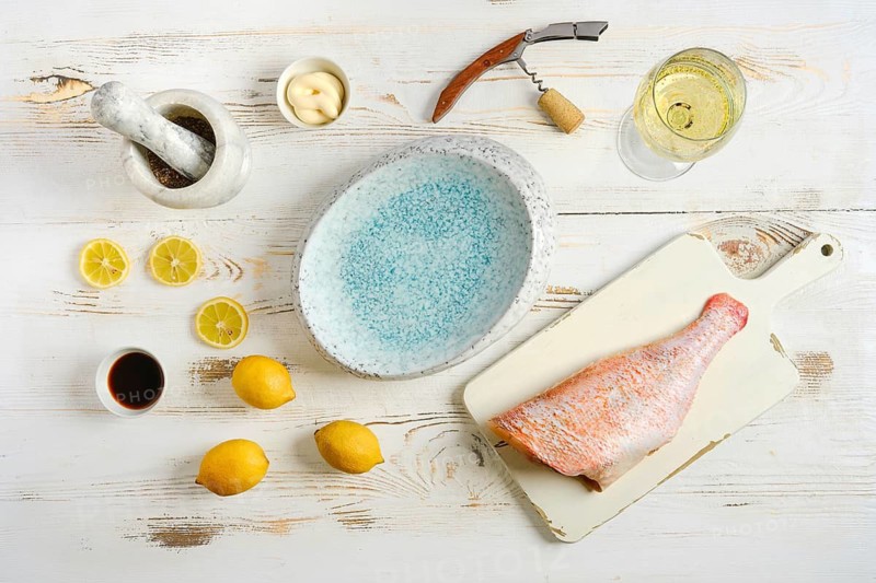 Red snapper lies on cutting board next to lemons, soy sauce, and mortar. Nearby, glass of white wine sits with corkscrew. Scene is set on wooden kitchen table in afternoon