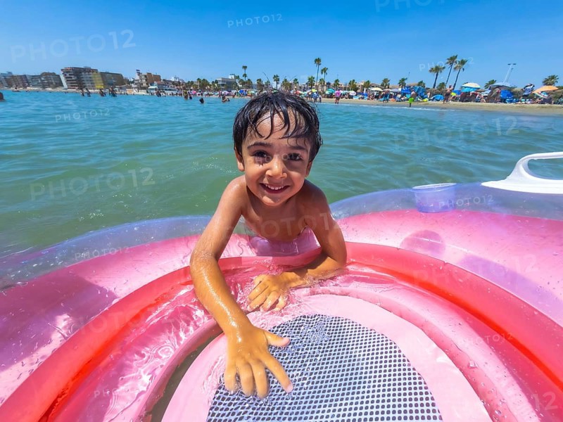 Adorable young boy smiling at the camera while playing in the ocean with an inflatable swimming ring, enjoying a sunny summer day at the crowded beach