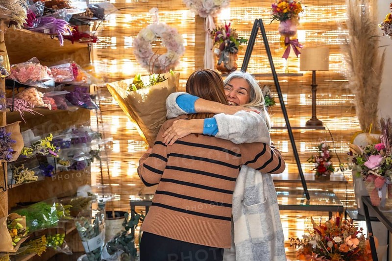 Caring daughter embracing her happy senior mother holding a bouquet of flowers in a cozy floral boutique, showing gratitude, love, and family connection