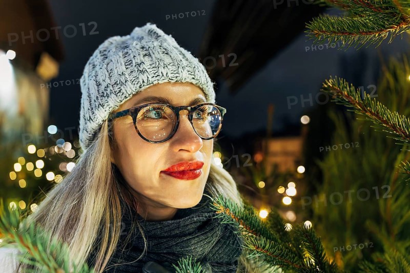 Young woman in glasses among fir branches and lights at night. A woman wearing glasses, a gray knit beanie and a dark scarf stands outdoors at night beside pine branches and warm glowing festive lights. Her long light hair and red lipstick catch the soft illumination, while blurred holiday bokeh fills the background. The close framing highlights winter textures, seasonal decorations and the cozy Christmas atmosphere