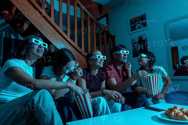 Family members sitting on a couch in a dimly lit living room, wearing 3d glasses, smiling and laughing while watching a film together and sharing popcorn
