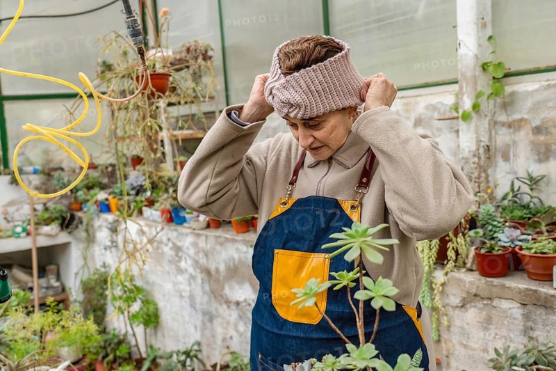 Older woman adjusts a pink knit headband on her head while wearing a denim apron inside her home greenhouse, surrounded by succulents and colorful potted plants