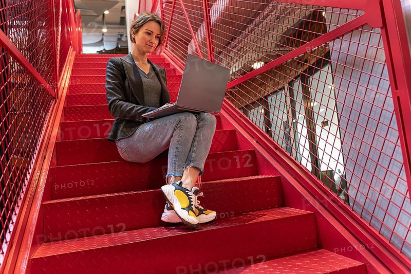Smiling young businesswoman working on laptop on red metal stairs in modern office