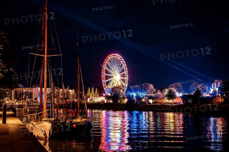Ferris wheel in neon light, evening at a funfair, seen from the waterfront, Lübeck, Germany