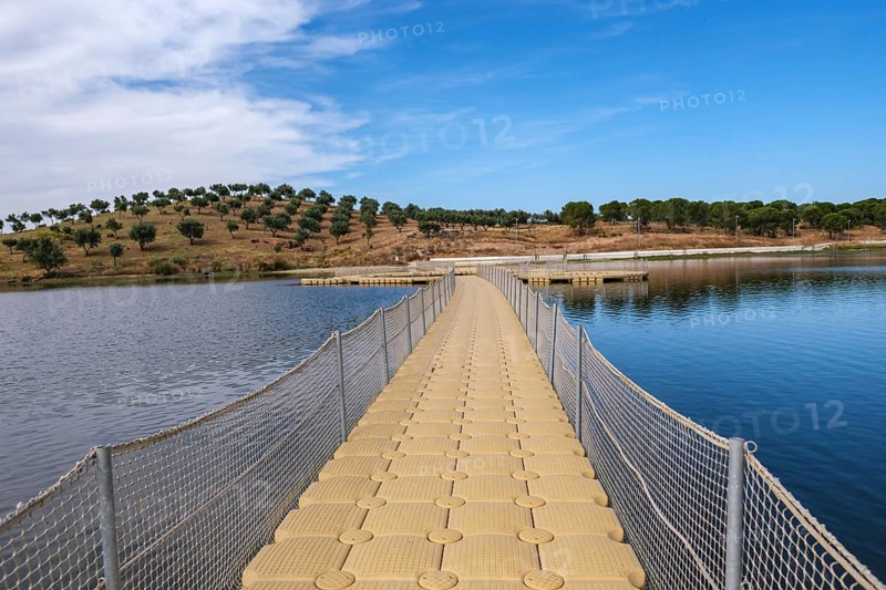 A floating jetty leads across calm water surrounded by green hills under a blue sky, Moura, Beja, Portugal
