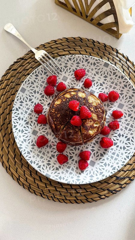 Stack of homemade pancakes topped with vibrant red raspberries, presented on a decorative plate over a woven placemat, offering a focus on healthy breakfast and brunch options