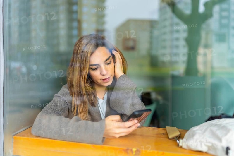 Woman sitting at a wooden cafe counter, absorbed in her smartphone while city reflections glide across the window glass, casual urban scene of modern quiet connectivity