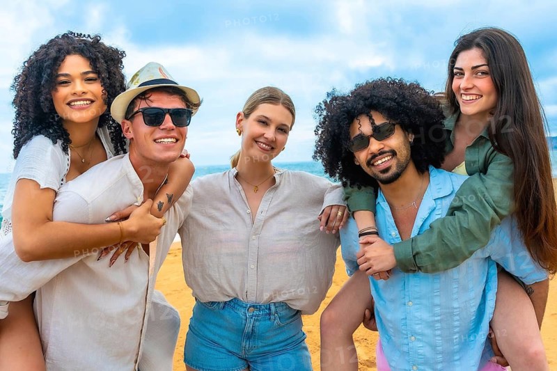 Diverse group of young adult friends smiling and enjoying summer vacation on a sandy beach under a blue sky, with two couples piggybacking, celebrating friendship and happiness