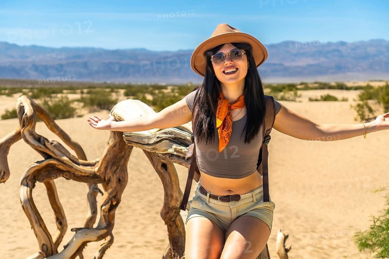 Young woman traveler posing happily on a piece of driftwood in death valley national park's mesquite flat sand dunes, appreciating the vast, arid desert environment under a clear blue sky