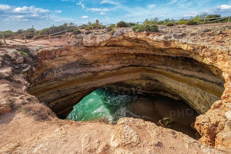Clifftop view into Benagil Cave in Algarve, Portugal, with a natural cave opening, layered limestone cliffs, Atlantic water, and rugged rock