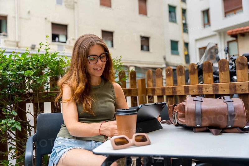 Young millennial woman wearing glasses and casual clothes working remotely on a digital tablet at an outdoor cafe, enjoying coffee and browsing online