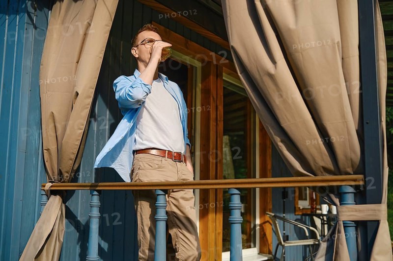 Middle-aged man stands at railing of patio terrace enjoying cold drink under hot afternoon sun