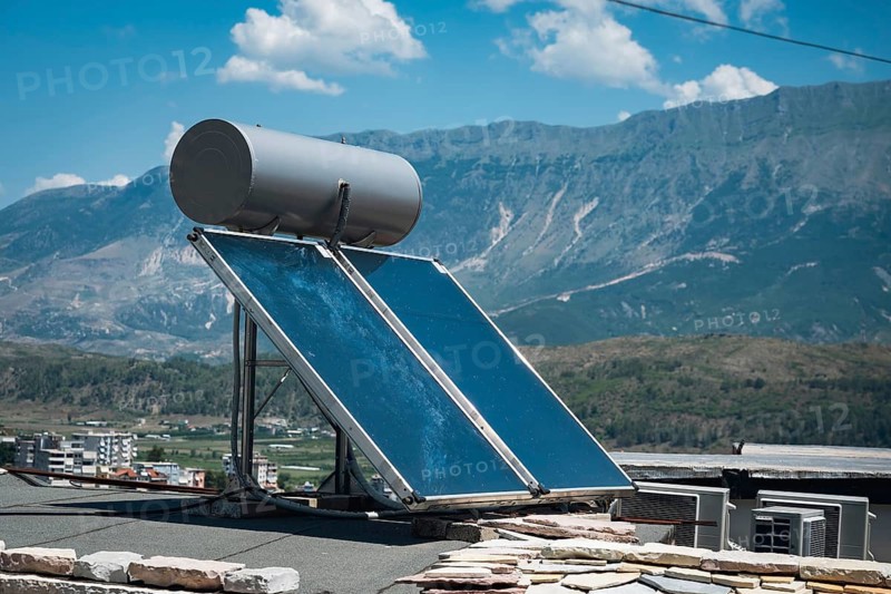 Solar water heating system and photovoltaic panels installed on rooftop with mountain landscape, showcasing renewable energy and sustainable technology under blue sky in Gjirokastra, Albania