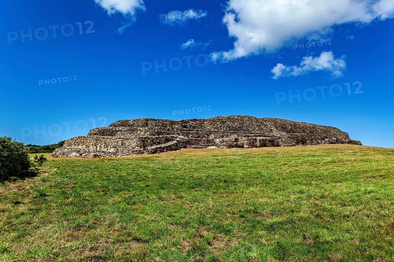 An old stone hill surrounded by a meadow under a clear blue sky with few clouds, The Cairn de Barnenez, Tomb of the Megalith Culture near Finistere in Brittany