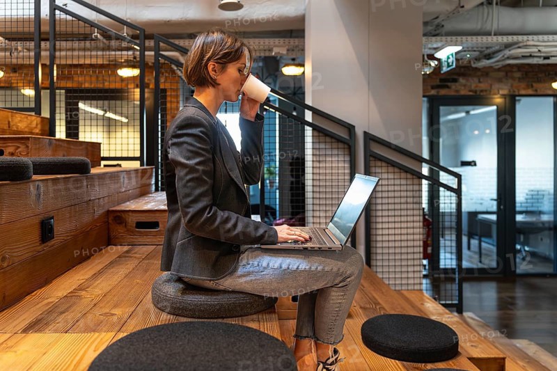 Young professional woman drinking from paper cup while working on laptop in coworking space