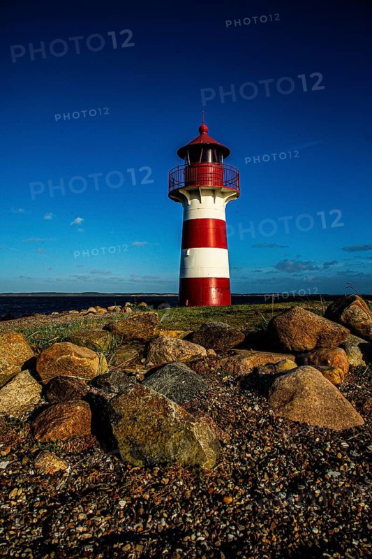 A red and white lighthouse stands under a clear blue sky on a rocky beach, Grisetaodde lighthouse, Neno Sound in the Limfjord, North Jutland, Denmark