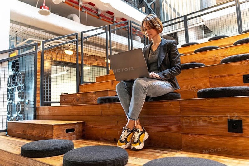 Young woman in jeans, blazer and sneakers working on laptop in modern coworking space with wooden steps