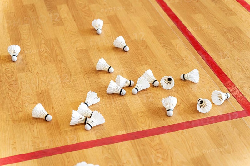 Several badminton shuttlecocks scattered across an indoor court with red boundary lines on a wooden floor. The scene reflects training or game preparation in a sports hall