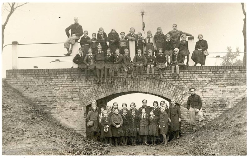 Girls, girls' class, trip, teacher, dark hiking clothes, large group of people standing and sitting on a bridge. Style is traditional, shot in black and white, around 1928