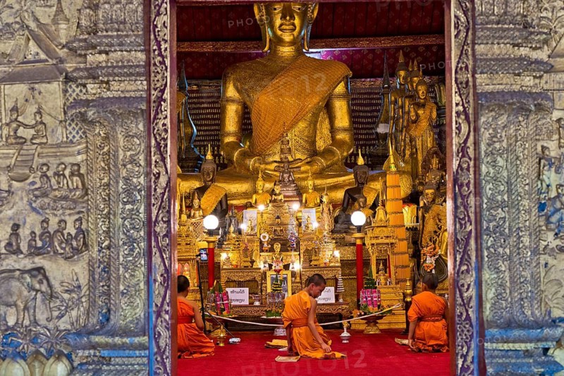 Buddhist monks and novices sit in front of a large Buddha statue in Wat May Souvannapoumaram, Luang Prabang, Laos, Southeast Asia