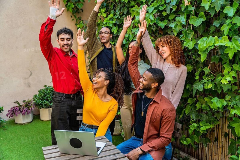 Cheerful diverse group of young adults raising hands in celebration, showing community support, teamwork, and achievement during a casual outdoor gathering or therapy session