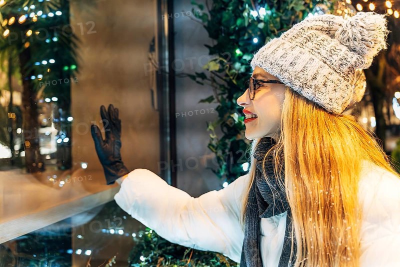 Smiling young woman in winter clothing looking at store window display. A woman wearing a white winter coat, gray knit hat with a pom-pom, black gloves and glasses stands beside a glass window decorated with warm holiday lights and greenery. Her gloved hand touches the reflective surface, capturing soft highlights from the surrounding illumination. The scene combines winter attire, glowing seasonal decorations and night reflections, creating a cozy festive atmosphere