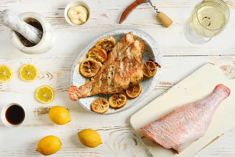 Grilled red snapper is presented on white dish with lemon slices and sauces nearby. Fresh lemons and mortar with spices sit around main dish on wooden table