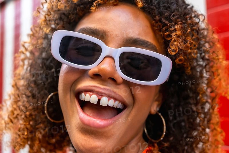 Young woman smiling widely, wearing fashionable white sunglasses and hoop earrings, presenting a cheerful and expressive face with diasteam, capturing a moment of joy and positive lifestyle outdoors