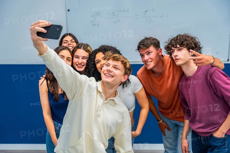 Happy multiethnic students gathering close together, smiling and posing for a group selfie in a classroom with a whiteboard showing mathematical equations
