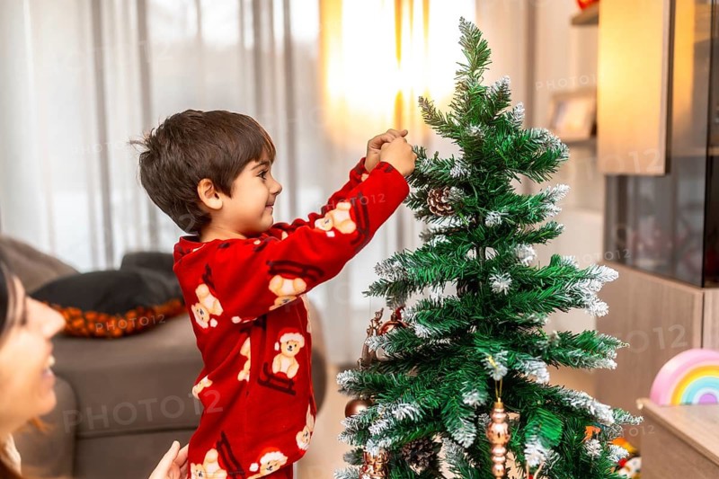 Young boy in red pajamas helps his mother hang ornaments and pinecones on the christmas tree, sharing a cozy, joyful holiday moment at home