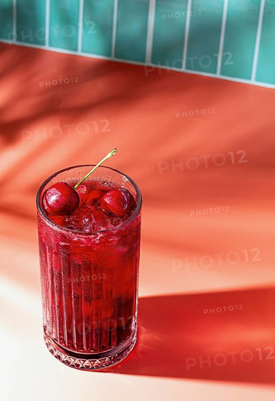 A glass filled with a cherry drink sits on a smooth surface. The drink contains ice and cherries on top. The background is bright red and well lit