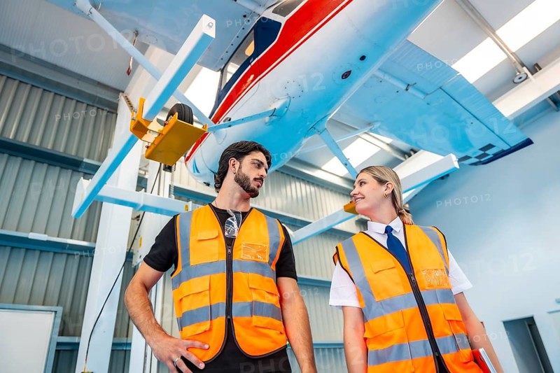 Two aviation professionals male mechanic and female pilot in high visibility vests stand in a clean hangar discussing inspection of a small airplane suspended overhead, teamwork focused