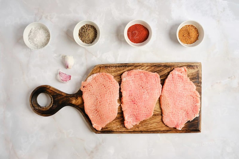 Three pieces of raw pork chops on wooden cutting board. Small bowls with spices sit nearby. Garlic cloves are placed next to the cutting board