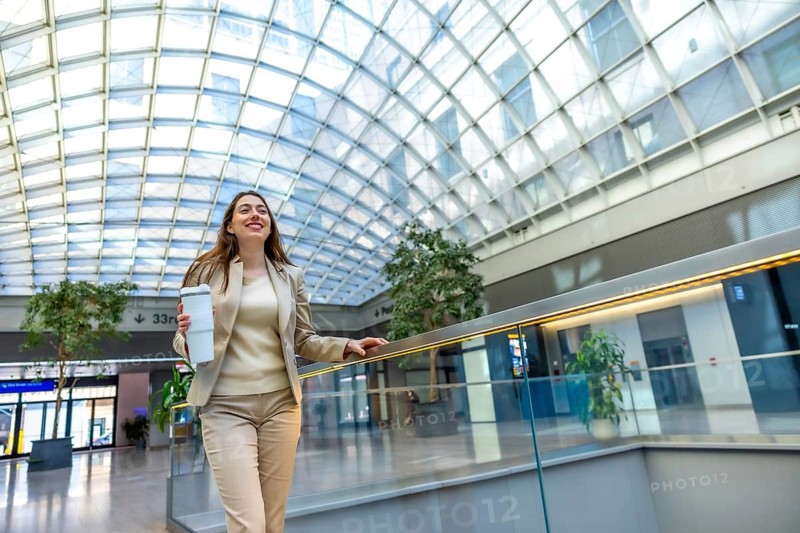 Young professional businesswoman walking confidently through a modern glass roofed transit hub, holding a reusable coffee cup and smiling as she heads toward her commute and career goals
