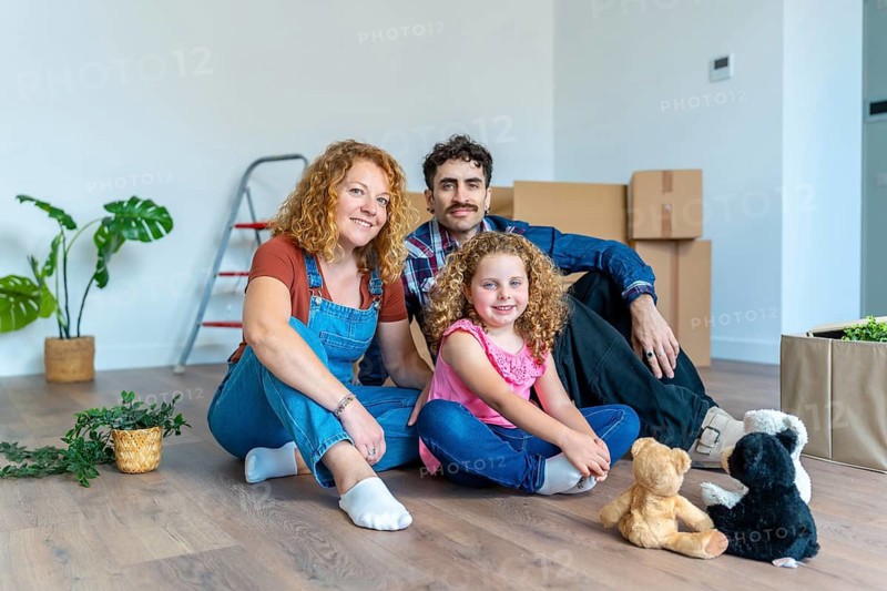 Cheerful family with a young daughter celebrating moving into their new house, sitting together on the floor surrounded by moving boxes and household items, anticipating a new life beginning