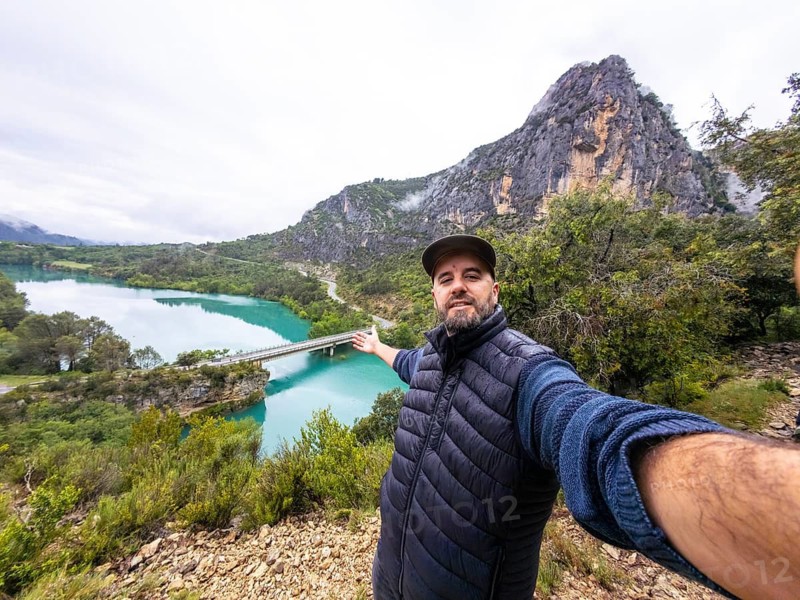 Man smiling at the camera taking a selfie, showing the entremon gorge landscape, featuring the vibrant turquoise cinca river, a bridge, and mountains in the pyrenees