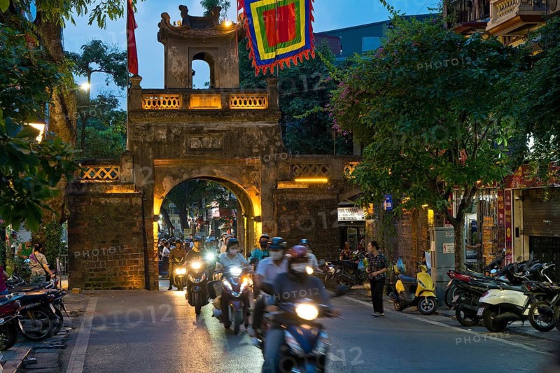 City gate during the evening rush hour on a busy street with mopeds, scooters, motorbikes and passers-by, surrounded by historic buildings and trees, Historic City Gate in the Old Quarter of Hanoi Vietnam