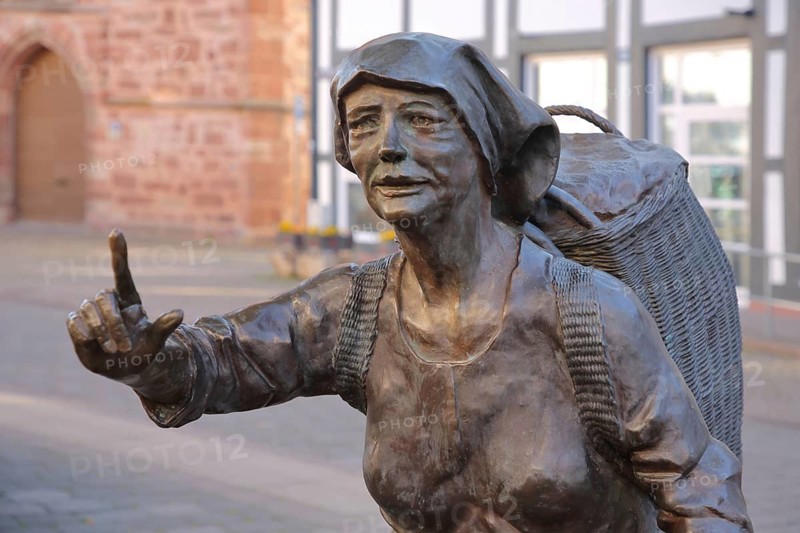 Market Women sculpture by Ewald Rumpf 2009, modern art, bronze sculpture, life-size, woman standing with index finger and wicker basket on her back, gesture, arm up, backpack, shopping basket, marketplace, Rotenburg an der Fulda, Hesse, Germany