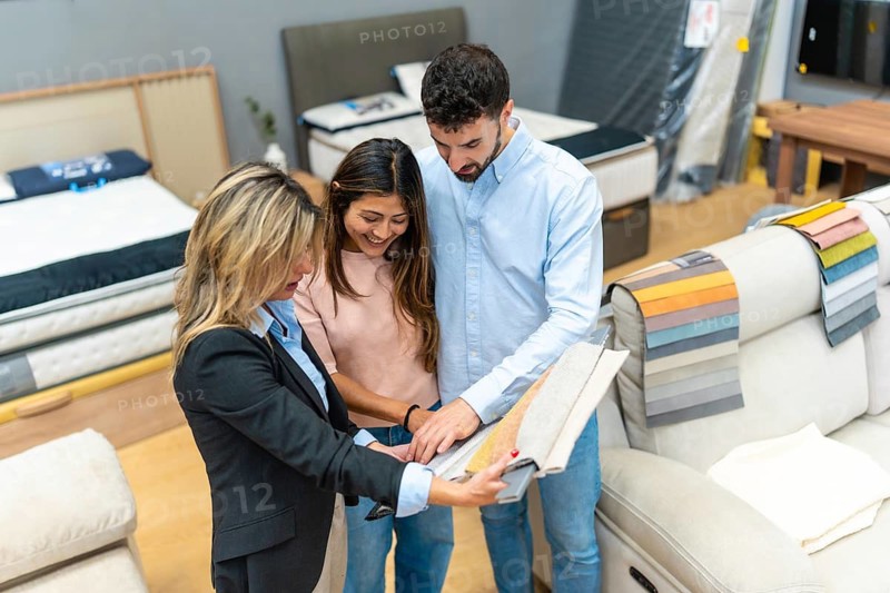 Sales assistant assisting a young couple in a home improvement store, showing different fabric swatches for furniture and discussing various design options for their new home