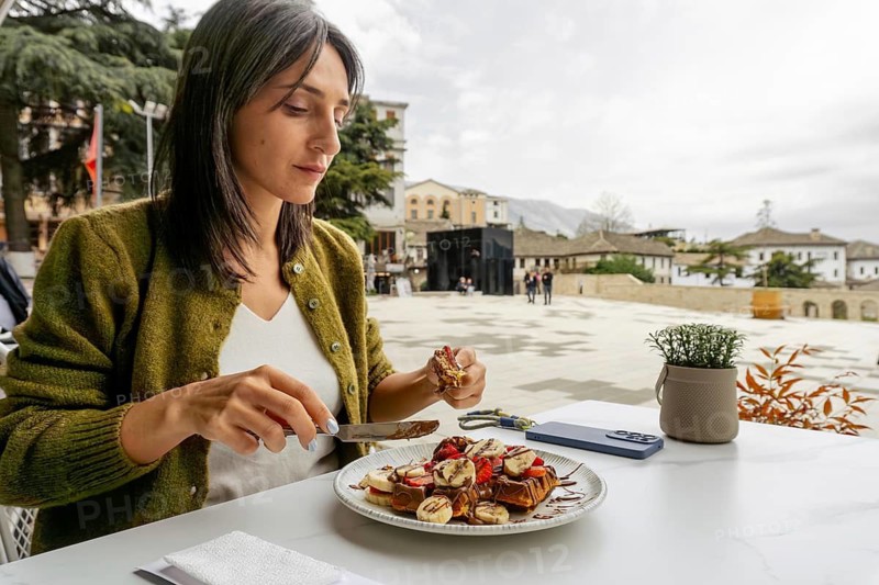 Close up woman sit in cafe enjoying large, indulgent waffle dessert portion topped with fruit and chocolate at an outdoor cafe. Travel lifestyle dining in historic stone town square of Gjirokastra