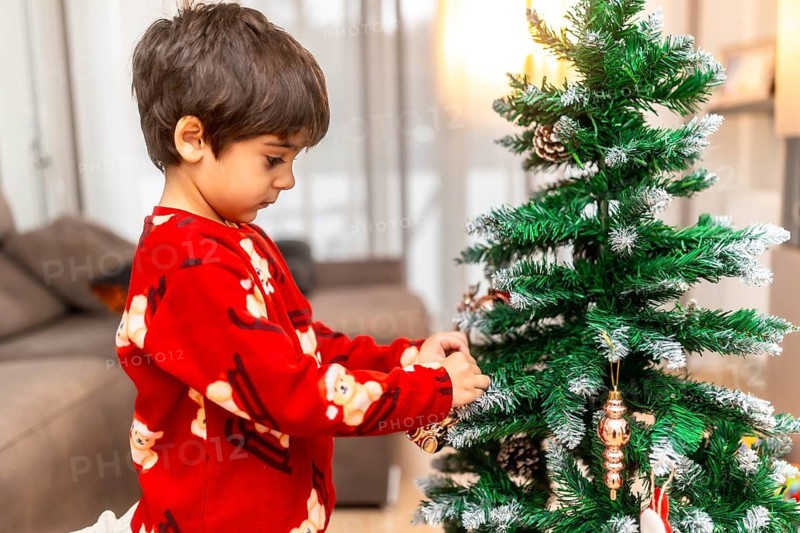 Happy child in red pajamas carefully hangs golden ornaments on a small christmas tree at home, creating a cozy, festive holiday scene filled with joy and tradition