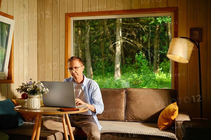 Middle-aged man sits at small round table with laptop in living room in wooden cabin. Sunlight enters through large window. Green trees are seen outside
