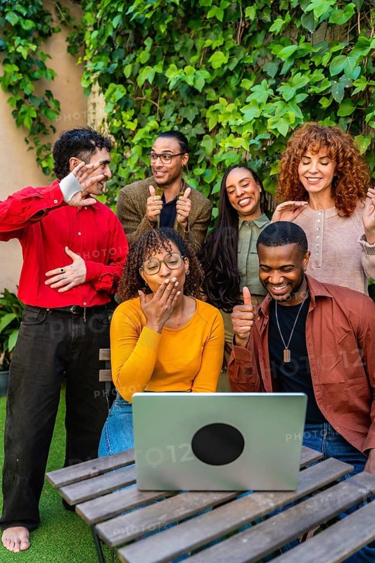 Diverse friends gathered on an outdoor patio, smiling and engaging in a supportive virtual group session via laptop, connecting, bonding, and sharing encouragement remotely