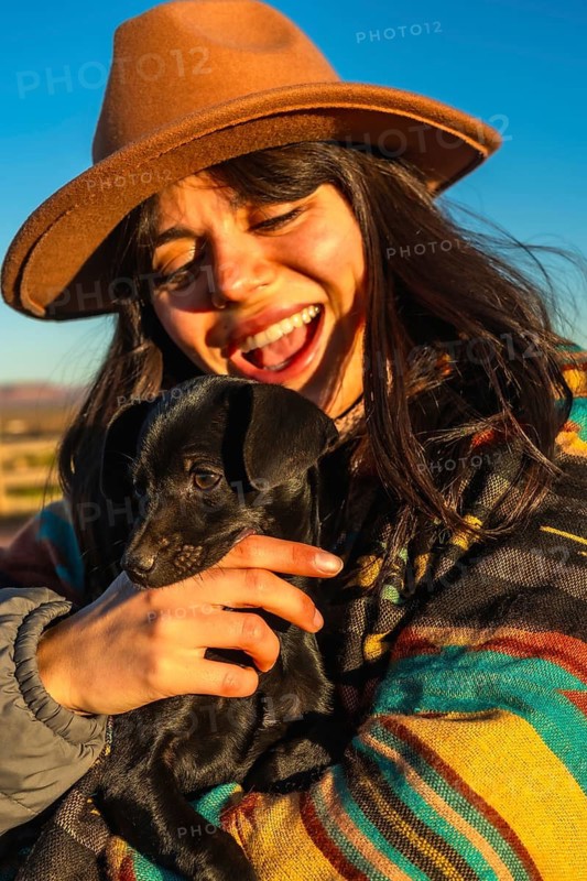 Woman in a brown hat and colorful poncho laughing and hugging a small black puppy at sunrise in monument valley, celebrating joyful outdoor bond and golden hour warmth