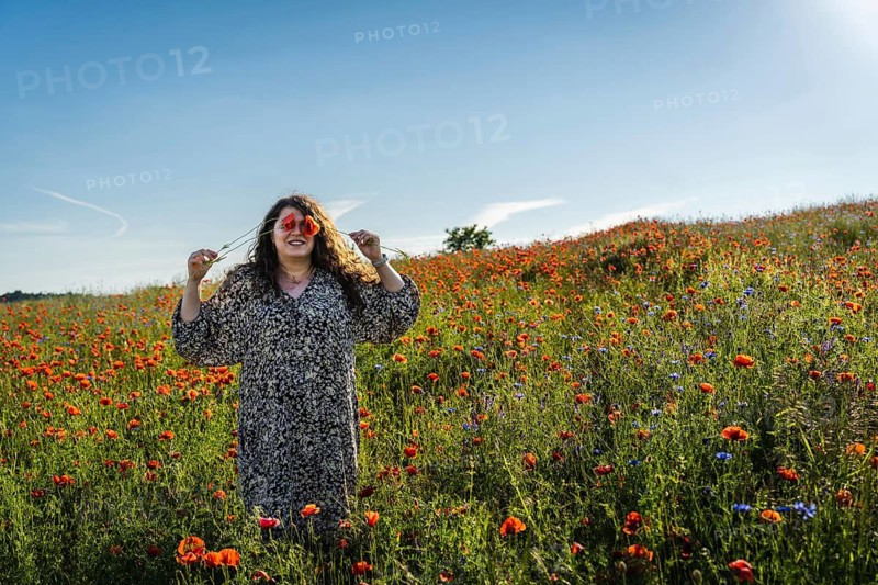 Plus size woman covering eyes with poppy (Papaver rhoeas) flowers while standing in blooming field under bright summer sun
